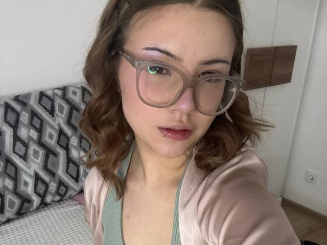 Close-up of BarrieGromley, a young woman with wavy brown hair, wearing large clear glasses and a pink hoodie over a green top. She gazes directly at the camera with a slight smirk. Her lips are painted dark red. The background includes a patterned black and white pillow and a wooden panel.