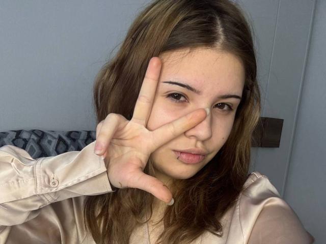 Fotografía de BarrieGromley, con piel clara y cabello castaño, haciendo el gesto de la paz con su mano derecha frente a su cara. Vestía una camisa blanca de seda, con los labios ligeramente entreabiertos. Tensión sexual evidente..
