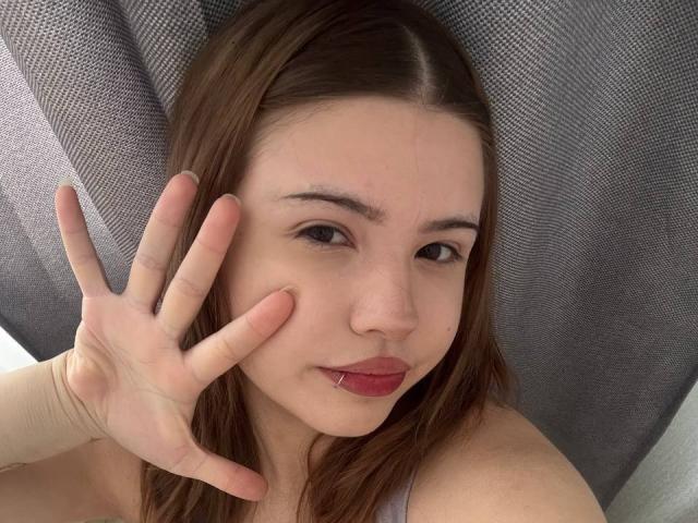 Close-up of BarrieGromley with brown hair, fair skin, and red lipstick, hand partially covering face, showing slight smile, relaxed expression, grey textured background, intimate and casual pose.