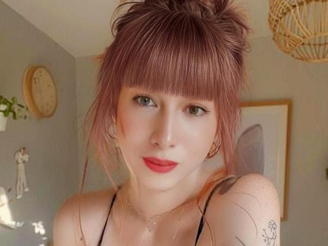 Primo piano di una SexyTopShemale con capelli rossi raccolti in uno chignon disordinato, frangia e rossetto rosso. Ha la pelle chiara, indossa un top nero a fascia, ha piccoli tatuaggi sulla spalla e uno sguardo sicuro. Lo sfondo include una lampada in vimini e un'opera d'arte a parete..
