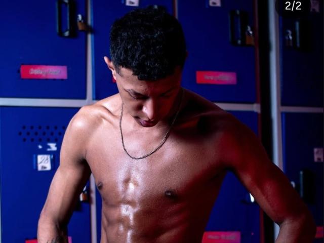 Fotografie von einem muskulösen JhonatanGuzman ohne Hemd, mit glänzender Haut, der eine Kettenkette trägt und steht. Vor blauen Schließfächern. Sein Penis ist nicht sichtbar. Die Zahl "21" befindet sich oben rechts..