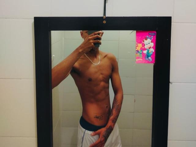 Fotografia di JhonatanGuzman, un uomo magro, senza camicia, con la pelle scura, addominali visibili e tatuaggi, che sta in piedi di fronte a uno specchio. Ha un telefono vicino al viso, indossa dei pantaloni bianchi. La sua mano è sulla parte inferiore del corpo..