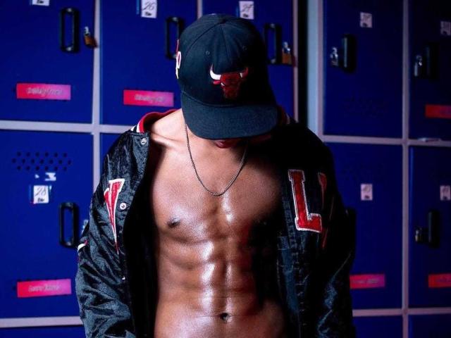 Fotografía de Jhonatan Guzman, musculoso, sin camisa, usando una gorra negra y una chaqueta negra con la letra "L" en rojo. Piel lisa y brillante, taquillas azules en el fondo. Abdominales prominentes, tensión sexual, complexión atlética..