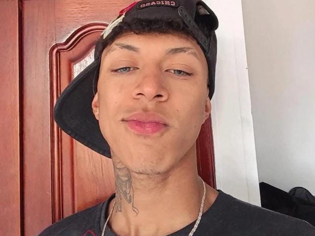 Close-up van JhonatanGuzman met een lichte grijns, die een zwarte pet en een ketting draagt, met een lichte huidskleur, een tatoeage op zijn nek, enigszins rommelig haar, rode deur op de achtergrond, casual stijl, subtiele aanwijzing van een erectie..