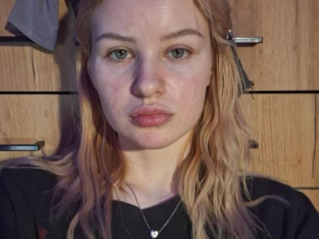 Close-up van RoseUty, een vrouw met bleke huid, blauwe ogen en roze lippen, die recht in de camera kijkt. Ze heeft blond, golvend haar en draagt een zwarte top met een ketting. Houten achtergrond.