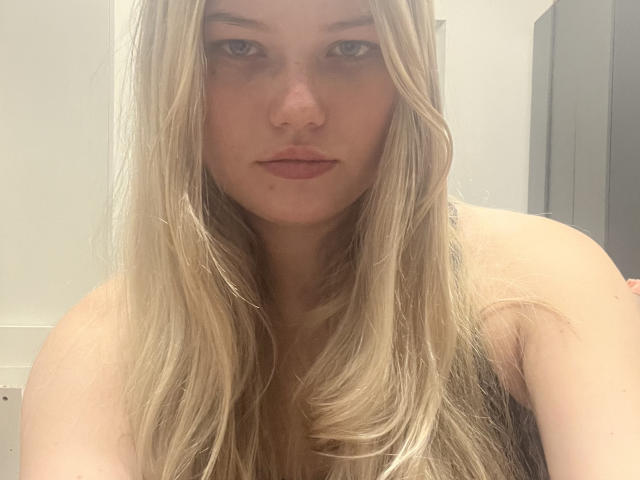 Close-up foto van RandaDaill, een blonde vrouw met lang, steil haar, die recht in de camera kijkt. Neutrale uitdrukking.
ze heeft bleke huid en draagt geen top, waardoor haar blote schouders te zien zijn. Een ongelooflijk intense blik..