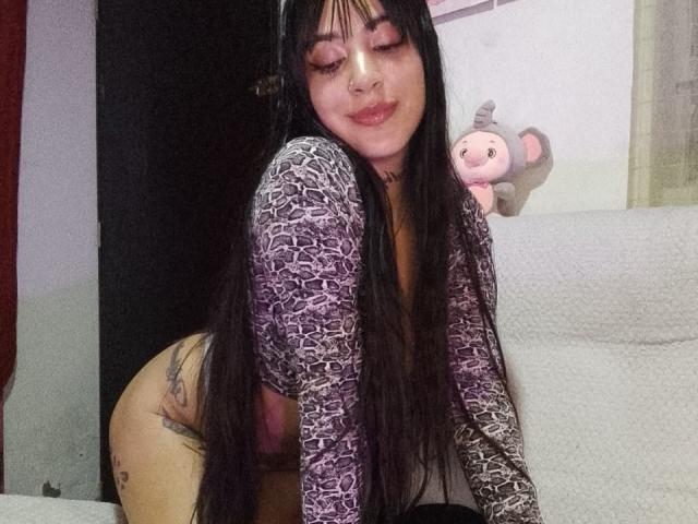 Fotografie von AnalisseJade mit langen, schwarzen Haaren, geschlossenen Augen und einem leichten Lächeln. Sie lehnt sich auf einer weißen Couch vorwärts, trägt ein gemustertes Oberteil und im Hintergrund ist ein Stofftier zu sehen..