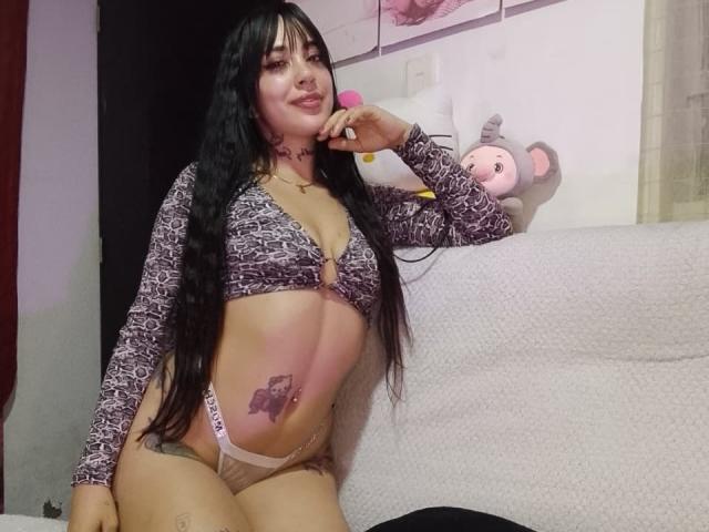 AnalisseJade, a curvy woman with long black hair, wears a patterned bra and pink panties. She leans on a bed, showing a belly tattoo, with a stuffed bear in the background.