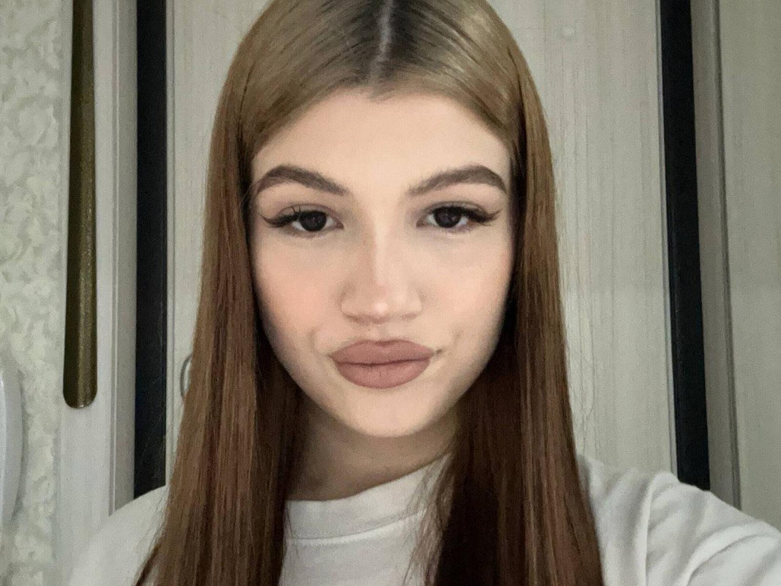 Primo piano di Jaclyn Cerasuolo, una giovane donna con lunghi capelli castani, labbra piene e sopracciglia folte. Indossa un maglione a collo alto bianco, e guarda direttamente la fotocamera con un sorriso appena accennato..