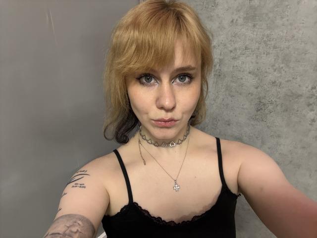 Fotografia de FenitaMolyn, uma jovem com cabelo castanho claro e franja, vestindo uma blusa de renda preta, um colar e colar.
ela tem tatuagens no braço esquerdo, olhando diretamente para a câmera com uma expressão séria..