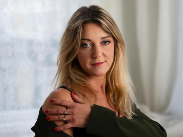 Photographie de MileyXStar, cheveux blonds, yeux bleus, haut noir à une épaule, ongles rouges, main sur l'épaule, fond blanc. éclairage doux, léger sourire, maquillage discret, alliance à l'annulaire, pose confiante, sexy..