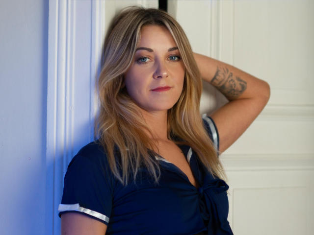 Foto van MileyXStar, staand voor een witte deur, leunend met één arm omhoog, waarbij een tatoeage op haar rechterarm zichtbaar is. Ze draagt een zwart shirt met witte accenten, heeft blauwe ogen en blond haar. Heel aantrekkelijk..