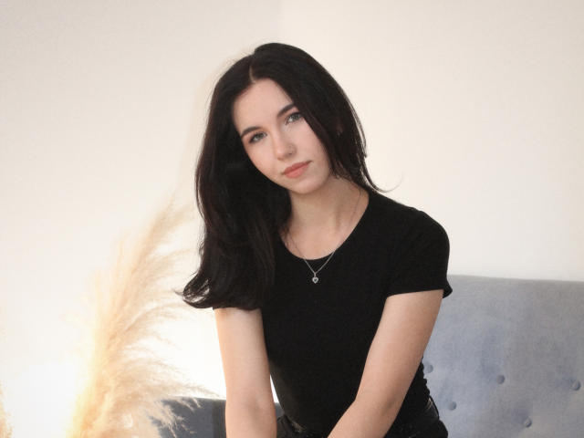 Photographie de Tricia Barnes, avec de longs cheveux noirs, portant un t-shirt noir, assise devant un fond blanc éclatant. Un coussin gris. Tension sexuelle, tellement mignon, léger sourire, lumière douce.