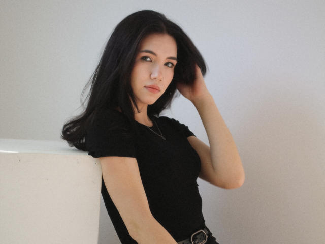 Tricia Barnes, con cabello negro largo, apoyada contra una superficie blanca, vestida con una blusa negra, con la mano en el cabello, mirada intensa. piel clara, ligera sombra en el lado derecho, pose sugestiva, iluminación suave..