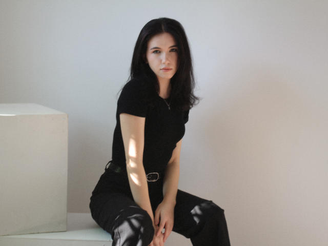 Tricia Barnes è seduta su un cubo bianco, con le gambe divaricate, indossando una maglietta nera e pantaloni neri. Ha i capelli lunghi e neri, la pelle chiara e un'espressione seria. Le ombre mettono in risalto le sue braccia..