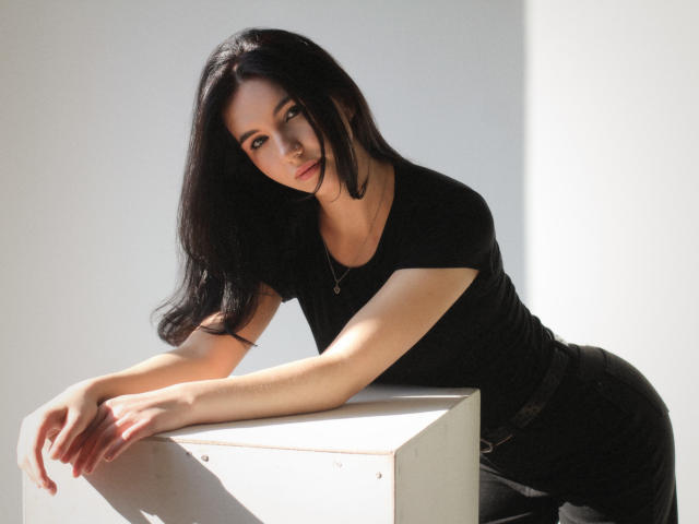 Tricia Barnes mit langem, schwarzem Haar, die nach vorne gebeugt auf einem weißen Würfel sitzt, trägt ein eng anliegendes schwarzes Hemd, und ihr Gesicht ist von hellem Licht beleuchtet. rechts, im Schatten liegend, sinnlicher Ausdruck, Licht, das ihre Kurven betont..