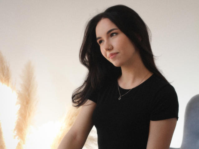 Tricia Barnes, mit langem, schwarzem Haar, trägt ein schwarzes T-Shirt und eine Halskette und blickt leicht zur Seite, mit einem subtilen lächeln. Der Hintergrund ist hell mit einem weichen Lichtschein. Verdammt sexy..