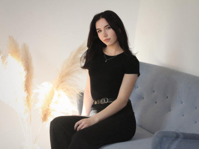 Tricia Barnes, con cabello largo y negro, vestida con una blusa y pantalones negros, sentada en un sofá azul con capitonado. La luz brillante detrás de ella crea un efecto de halo, resaltando su expresión seria. Increíblemente atractivo/a..