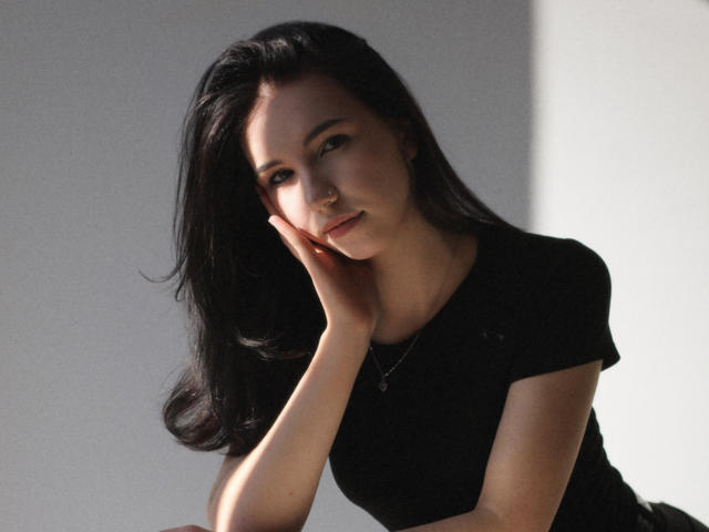 Fotografia di Tricia Barnes, una donna dai capelli scuri con un'espressione seducente, appoggiata a una mano, indossa una maglietta nera, con ombre delicate. luce intensa a destra, alto contrasto, sguardo intenso, sexy, posa provocante.