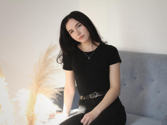 Tricia Barnes sitzt auf einem grauen Sofa und trägt ein schwarzes T-Shirt und eine schwarze Hose mit einem braunen Gürtel. Sie hat langes, schwarzes Haar, blasse Haut und einen ernsten Gesichtsausdruck. Verdammt heiß..