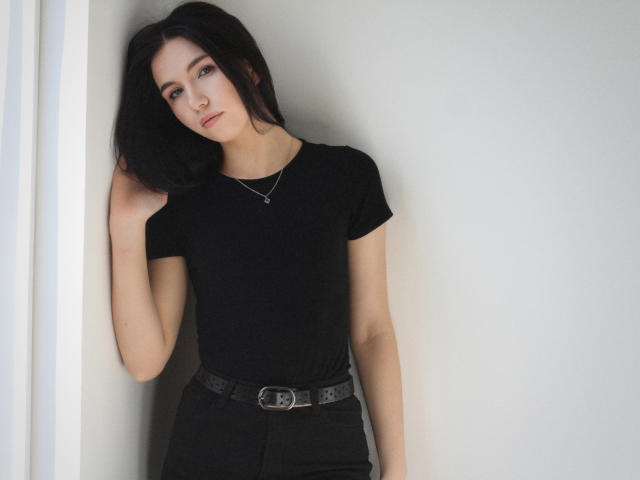 TriciaBarnes stands against a white wall, leaning slightly to the left with one hand in her black hair. She wears a tight black t-shirt and black pants, belted with a silver buckle. She's looking directly at the camera with a slight, sexy smirk.