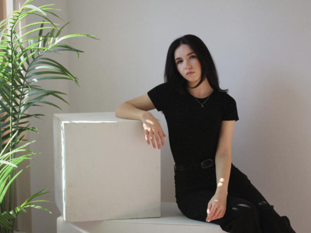 Tricia Barnes, com longos cabelos pretos, sentada no chão encostada em um cubo branco, vestindo uma camiseta preta e calças, inclinada. no cubo com o braço, a planta à esquerda. Muito atraente..