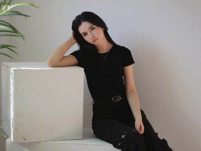 Tricia Barnes, com pele clara, cabelo preto e roupa preta, apoiada em um cubo branco, com a mão atrás da cabeça, e uma planta verde ao lado. canto esquerdo, sentada, sexy, confiante, minimalista, luz suave.