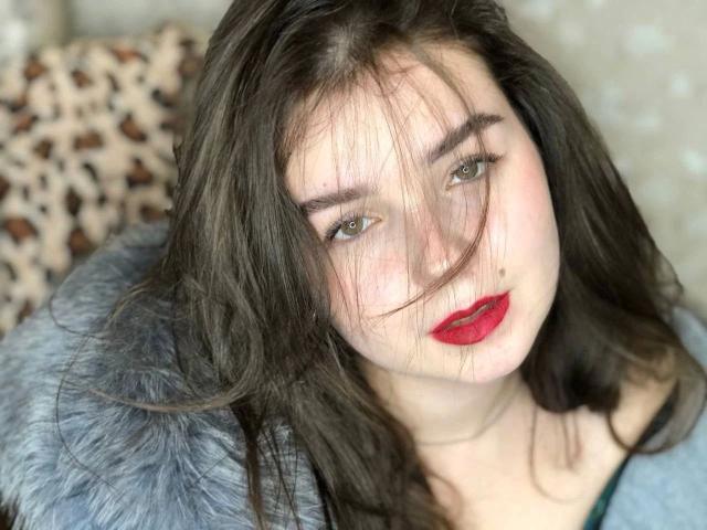 Fotografia ravvicinata di SofyX-hot con pelle chiara, capelli neri e rossetto rosso, che indossa un cappuccio di pelliccia grigia. Leggere ombre sul suo viso, occhi verdi intensi e uno sguardo seducente..