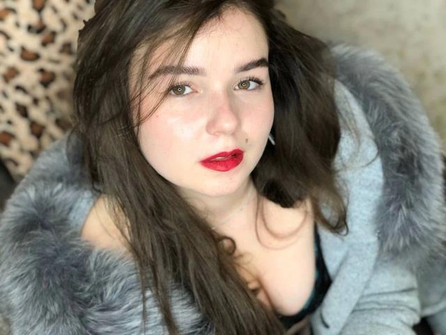 Fotografia ravvicinata di SofyX-hot con pelle chiara, rossetto rosso e capelli castano scuro. Indossa un cappotto di pelliccia grigia e guarda verso l'alto con occhi verdi seducenti. Sfondo con motivo leopardo. Incredibilmente sexy..