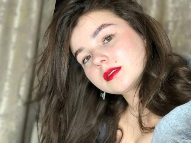 Primo piano di SofyX-hot con lunghi capelli scuri, pelle chiara e rossetto rosso acceso. Lei guarda in modo seducente, con la testa inclinata, indossa una maglietta grigia, lo sfondo è sfocato. I suoi occhi e le sue labbra sono i punti focali..