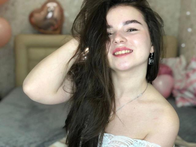 SofyX, met lang zwart haar, lachend, hand in haar, draagt een witte kanten top zonder bandjes, felle rode lipstick, oorbellen. Hel licht op het gezicht, zachte achtergrond met een hartvormig object en ballonnen..