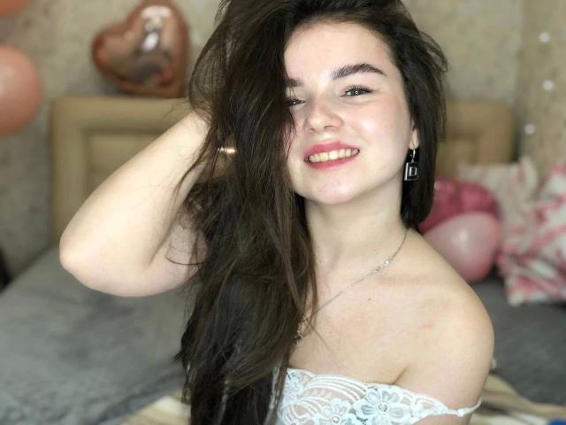 SofyX, mit langen, schwarzen Haaren, lächelnd, eine Hand im Haar, trägt eine weiße Spitzenbluse, blasse Haut, kleine Brüste, Ohrringe. Unschärfer Hintergrund mit herzförmiger Dekoration und rosa Ballons. Offensichtliche sexuelle Anspannung..