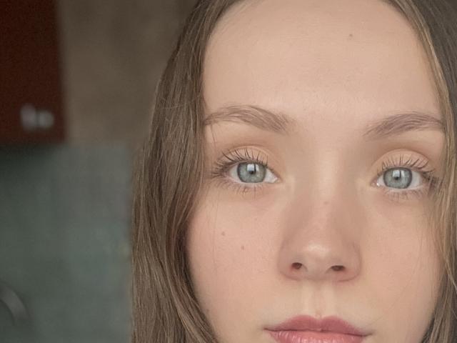 Photographie en gros plan de MirellaBagne, montrant sa peau pâle, ses yeux bleus et ses cheveux bruns. Son expression est neutre, avec des lèvres légèrement entrouvertes et rosées. L'arrière-plan est flou, ce qui met en valeur son visage. Regard extrêmement intense..