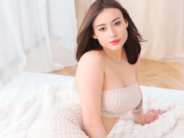 Photograph of KaylaDirtyLove, a brunette woman with red lipstick, leaning forward on a white bed, wearing a strapless white dress, showing cleavage, soft lighting, wooden floor in background.