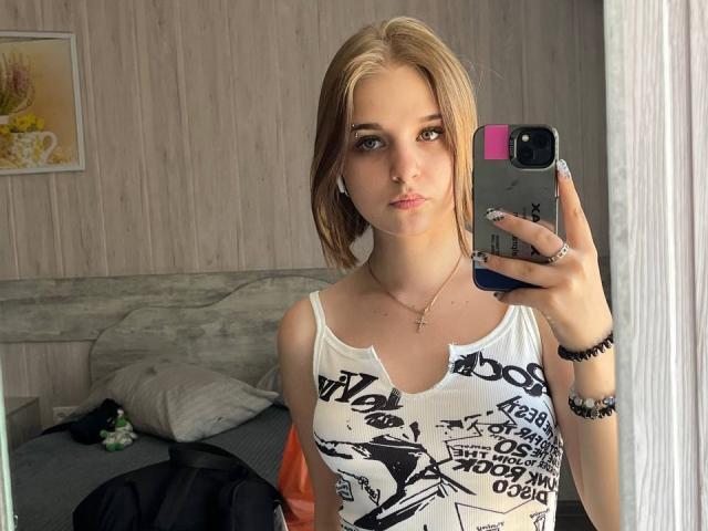 Fotografia de WowMonkey tirando uma selfie, segurando um celular preto com uma capa rosa, vestindo uma camiseta branca com detalhes pretos. Impressão gráfica, cabelo castanho claro, expressão neutra. Ambiente de quarto..