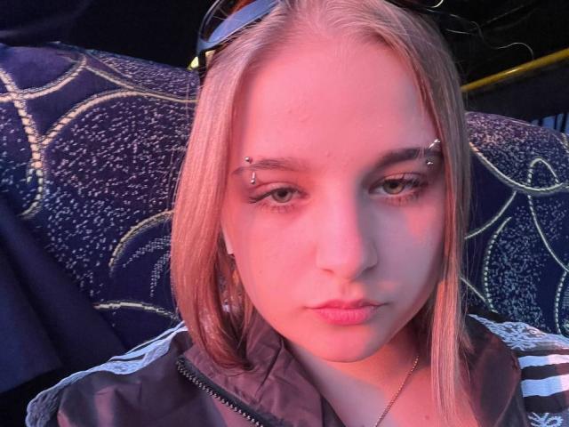 Close-up photo of WowMonkey, a blonde woman with piercing in both eyes, wearing a black and white jacket. Neon pink and blue lighting emphasizes her face and piercing. Fucking intense gaze.