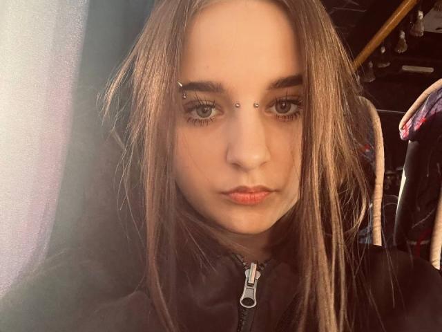 Close-up of WowMonkey, a young woman with straight brown hair, wearing a black jacket with a silver zipper. She has facial piercings, including a nose and upper lip ring. Serious expression.