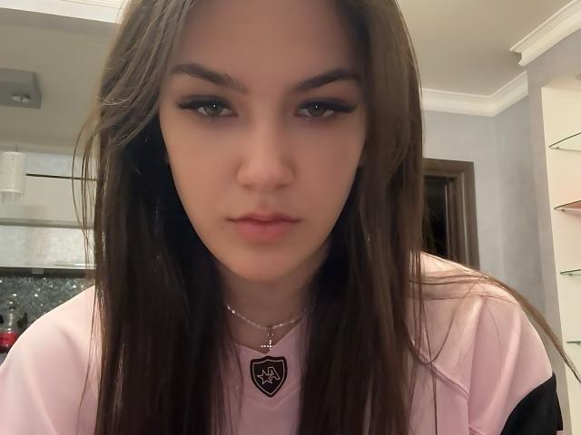 Gros plan de HyoRiopel, avec de longs cheveux bruns, une peau pâle et un eye-liner noir. Elle porte un haut rose avec un collier en forme de cœur, et regarde directement l'appareil photo avec une expression sérieuse..
