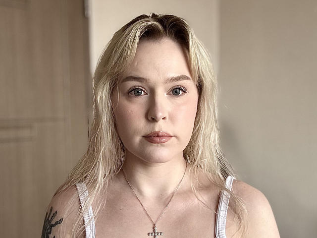 Fotografia di Maryan Batteen, al centro, con capelli biondi umidi, pelle chiara e un'espressione seria. Indossa una canotta bianca, una piccola croce come ciondolo e ha un tatuaggio sulla spalla sinistra. Luce intensa, sfondo neutro
.