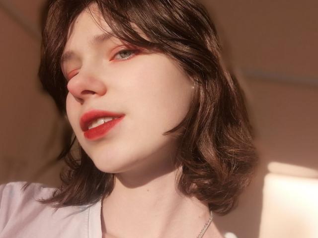 Close-up foto van NessaV, met een lichte huid, donkerbruin, golvend haar, felrode lipstick, een lichte glimlach, en zonlicht. Verlicht gezicht, zachte schaduwen, subtiele make-up, zichtbare ketting, lichte erotische spanning, verleidelijke blik..