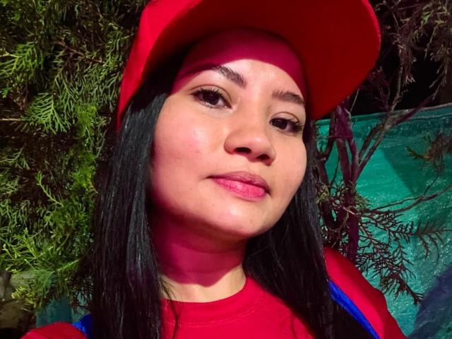 Primo piano di KatyViper, una giovane donna con lunghi capelli neri, che indossa un berretto rosso e una maglietta rossa. Ha una carnagione marrone chiaro, labbra rosa e un'espressione leggermente sensuale. Verde sullo sfondo.