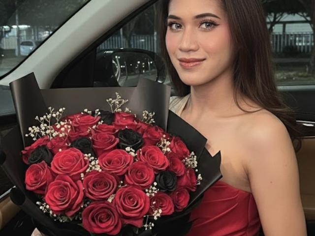 PreciousHorny, una mujer de piel clara con cabello largo y castaño, vestida con un vestido rojo sin tirantes, sostiene un ramo de flores rojas y rosas negras con espigas blancas, colocadas en un coche..