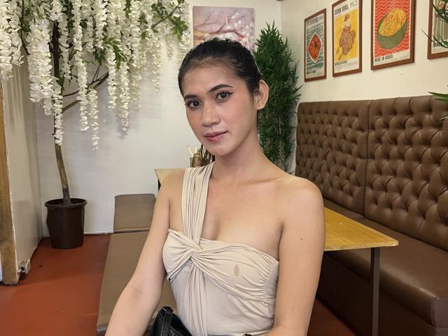 PreciousHorny with dark hair, tied back, wearing a strapless white top with diagonal stripes, sitting at a brown leather booth in a restaurant. White flowers hang above, colorful posters on the wall.