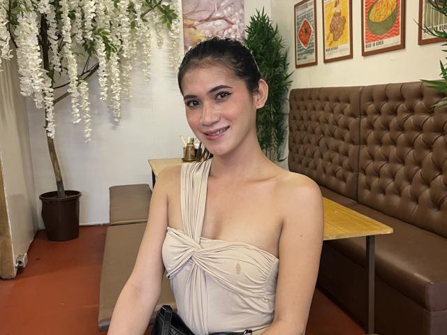 PreciousHorny sits in a brown tufted booth, wearing a white strapless top, smiling with a hint of arousal. Hanging white flowers, potted plant, and colorful posters in the background. Fucking sexy.