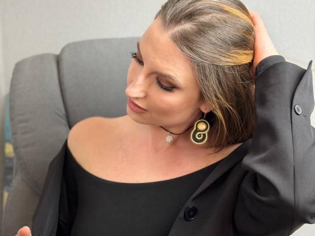 Milagres, with shoulder-length brown hair, leans back in a gray chair, hand in hair, wearing a black off-shoulder top, black jacket, and large spiral earrings, exuding relaxed, sexy vibes.