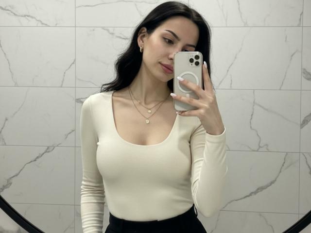 Valnes with long black hair takes a selfie in a marble-tiled bathroom. She wears a tight white long-sleeve top, showing her large breasts, and holds a white iPhone. Her expression is confident.
