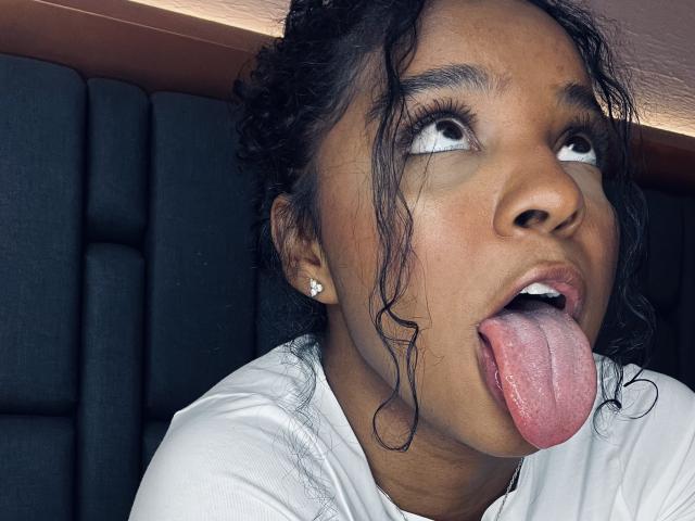 Foto em close-up de LisaMendez, com pele escura, cabelo cacheado molhado e olhos grandes, com a língua rosa comprida para fora. Ela está vestindo uma camisa branca e brincos de diamantes, olhando para cima..