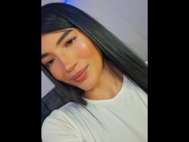 Nahaufnahme von PaulethJoens, einer jungen Frau mit langen, schwarzen Haaren, die ein weißes, geripptes Hemd trägt. Sie hat glatte Haut, volle Lippen und ein leichtes Lächeln. Ihr Penis ist nicht sichtbar..