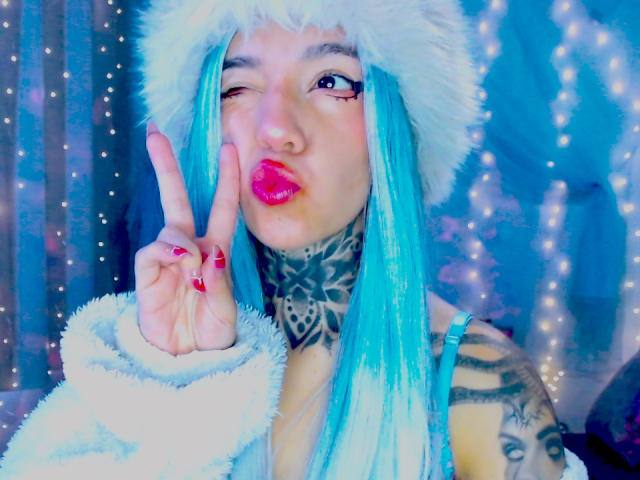 LannieGonzales, a cute Asian woman with blue hair, winks and sticks out her tongue while flashing a peace sign. Wearing a white fur hat and jacket, blue hair, red nails.