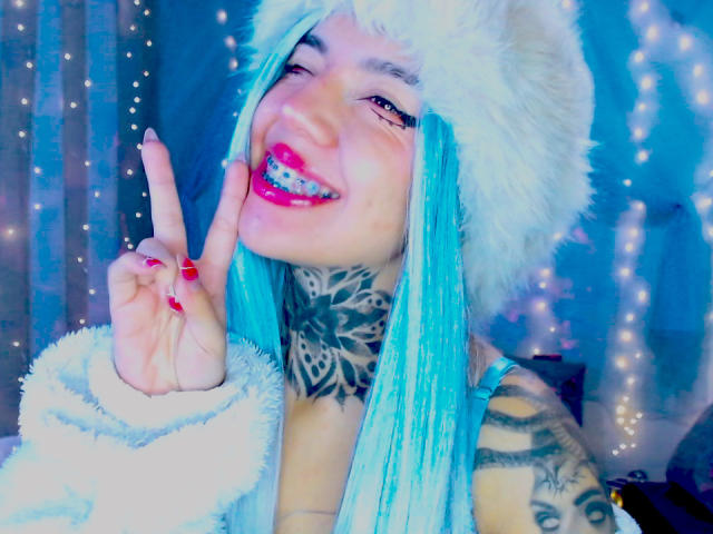 Nahaufnahme von LannieGonzales, die lächelt und die Zunge herausstreckt, mit blauen Haaren, einem weißen Fellhut, einem Finger, der nach oben zeigt, roten Nägeln und einem aufwendigen Outfit. Tattoo am Hals, blauer Streifen-Sleeve, glitzernder blauer Hintergrund, sexy, verspielt, intensiver Blick..